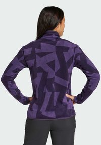 Chaqueta de forro polar púrpura con un patrón geométrico en tonos más oscuros de púrpura, con un cuello alto y mangas largas. Textura suave, ajuste casual.