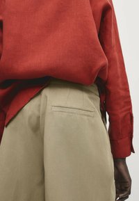 Chemise en lin rouge avec manches retroussées, associée à un pantalon beige doté d'une poche arrière détaillée. Le tissu semble texturé et respirant.