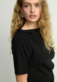 Černý top s krátkými rukávy, hladká textura a lodičkový výstřih. Modelka má vlnité blond vlasy a nosí velké stříbrné náušnice ve tvaru kruhu.