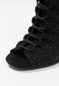 Zwarte suède peep-toe laars met een vetersluiting aan de voorkant, een gestructureerd oppervlak en metalen versieringen aan de zijkant.