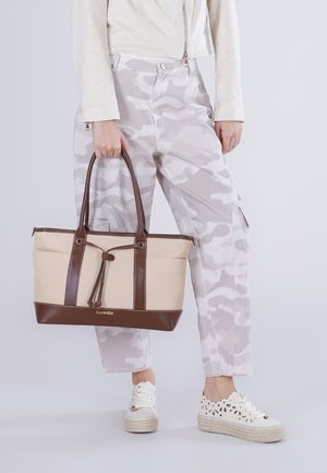 Osoba v světle béžových maskovacích kalhotách, krémové bundě, bílých perforovaných teniskách, držící béžovou a hnědou tašku typu tote s koženými rukojeťmi.