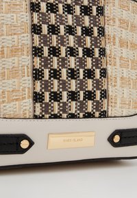 Sac à main tissé avec un motif à carreaux beige et noir, ferrures dorées, matériau texturé, avec une plaque de marque et des accents contrastants.