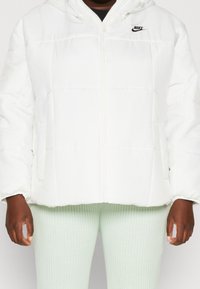 White puffer jacket with a zipper and hood. Features a quilted design and a small black Nike logo on the left chest area.