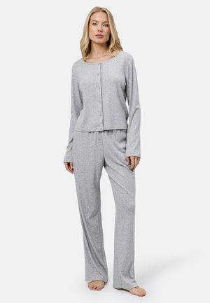 Kobieta stoi boso, ubrana w jasnoszary, długi rękaw z guzikami i dopasowane luźne spodnie na jednolitym jasnym tle.