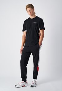 Black short-sleeve t-shirt with white logo, paired with black sweatpants featuring red and gray side panels, and white athletic shoes.