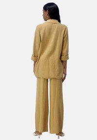 Completo in tessuto giallo chiaro e strutturato, composto da una camicia lunga con bottoni e maniche arrotolate, e pantaloni a gamba larga, entrambi con vestibilità rilassata.