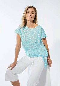 Hellblaues Oberteil mit einem dezenten weißen abstrakten Muster, kurzen Ärmeln und einem Seitenbindedetail. Getragen mit weißen, knöchellangen Hosen.