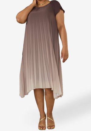 Donna che indossa un vestito ombré plissettato a maniche corte e sandali dorati con cinturini, in piedi su uno sfondo semplice.