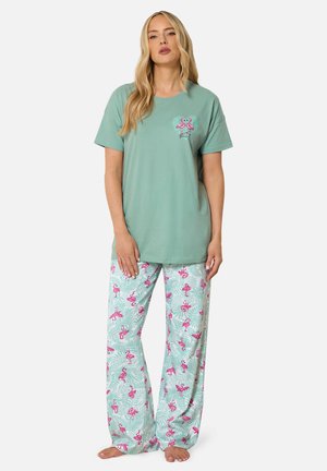 Femme portant un haut vert clair à manches courtes avec de petits flamants roses et un pantalon clair assorti avec un imprimé flamant rose, debout pieds nus.