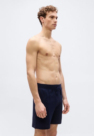 Νέος άντρας με σγουρά μαλλιά στέκεται γυμνόστηθος φορώντας σκούρα μπλε πλεκτά σορτς, πάνω σε απλό φωτεινό φόντο.