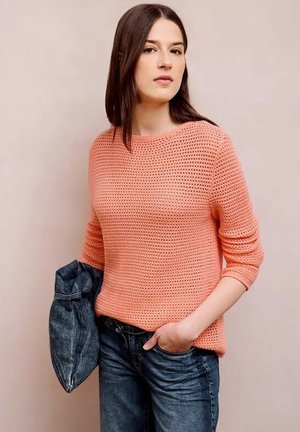 Nainen, jolla on suorat tummat hiukset ja päällään korallinvärinen neulepusero ja siniset farkut, pitäen toisessa kädessään taiteltua farkkutakkia.