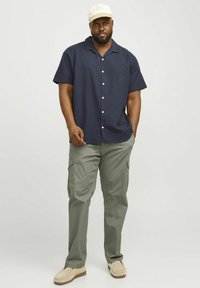 Navy strukturiertes Kurzarmhemd mit weißen Knöpfen, kombiniert mit olivfarbenen Cargo-Hosen. Das Model trägt eine hellfarbige Mütze und beiger Slip-On-Schuhe.
