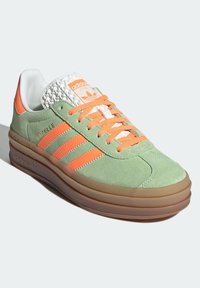 Sneaker in suede verde chiaro con accenti arancioni, tre strisce e una linguetta bianca testurizzata. Presenta una suola in gomma con una leggera elevazione.