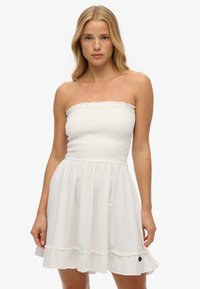Biała sukienka bez ramiączek z marszczonym gorsetem, rozkloszowanym dołem i falbaną na końcu. Wykonana z lekkiego materiału, z małym czarnym akcentem na dolnej krawędzi.