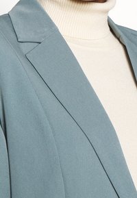 Vaaleansininen bleiseri terävillä läpällä vaalean kermanvärisen turtleneckin päällä. Pehmeä pinta, räätälöity muotoilu ja siistit linjat.