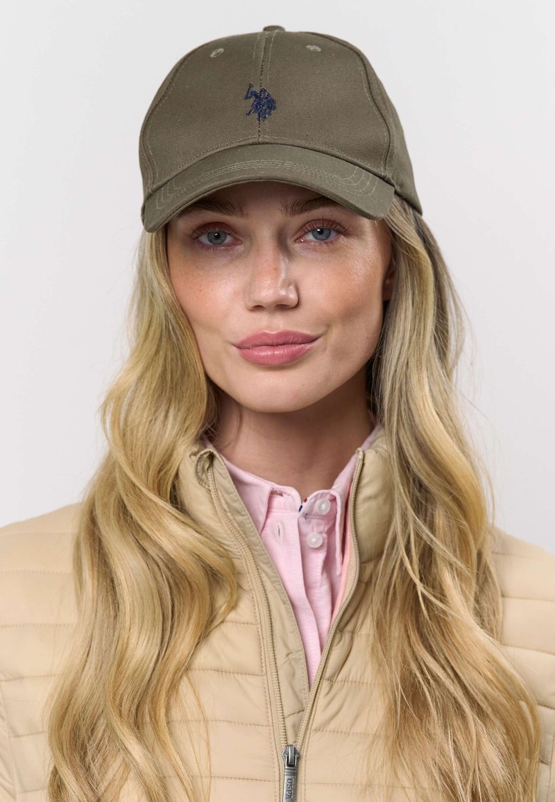 Gorra verde oliva con un logo bordado azul, combinada con una chaqueta acolchada beige sobre una camisa rosa claro con botones. Materiales con textura visibles.