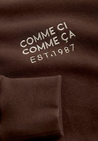 Hnedá tkanina s ryhovanou textúrou, na ktorej je vyšívaný text: "COMME CI COMME ÇA" a "EST. 1987" v svetlej farbe. Viditeľný detail manžety.