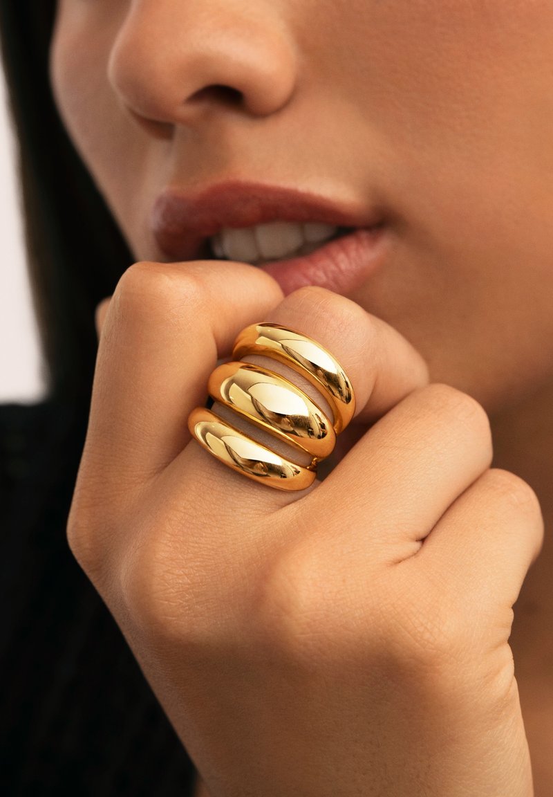 Anillos de metal en tono dorado con un acabado suave y brillante, que presentan tres bandas interconectadas en un diseño redondeado sobre una mano.