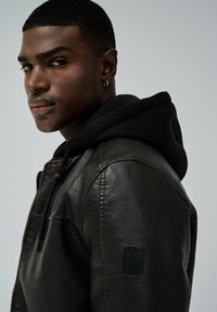 Bruine leren jas met een zwart hoodie sweatshirt eronder. De jas heeft zichtbare stiksels en een label met de tekst "SALSA JEANS."