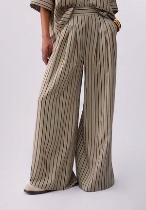 Pantaloni a gamba larga con righe verticali marroni e bianche, indossati con top abbinato a righe e scarpe beige a punta.