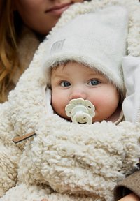 Krämsfärgad fleeceoverall med texturerat, fluffigt material; baby med blå ögon och en blommig napp, klädd i en ljusstickad mössa.