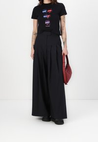 Černé tričko s barevným grafickým designem obličejů s brýlemi. Vysoké, plisované černé kalhoty, červená kabelka, černé boty.