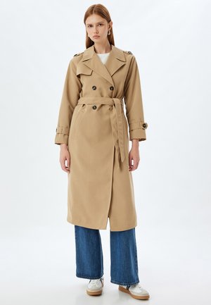 Manteau beige en trench avec des boutons double boutonnage, une ceinture et des poches latérales. Associé à un jean bleu à jambes larges et des baskets blanches.