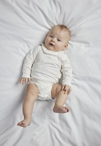 Beba u svijetlosmeđem dugom bodiju s ruffles ovratnikom, odmara na bijelom platnu, s kratkom kosom i plavim očima.