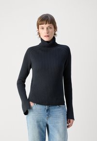 Maglione nero a collo alto a coste con un design aderente e maniche lunghe, abbinato a jeans di denim blu. Sfondo neutro.