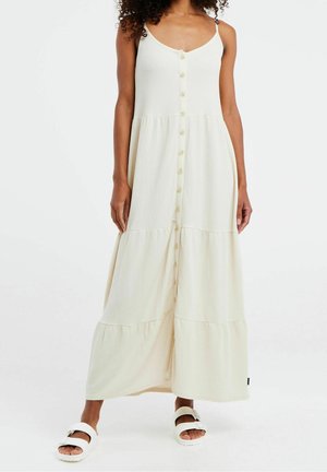 Femme portant une robe longue crème sans manches à boutons avec jupe à volants et sandales blanches, debout devant un fond blanc.