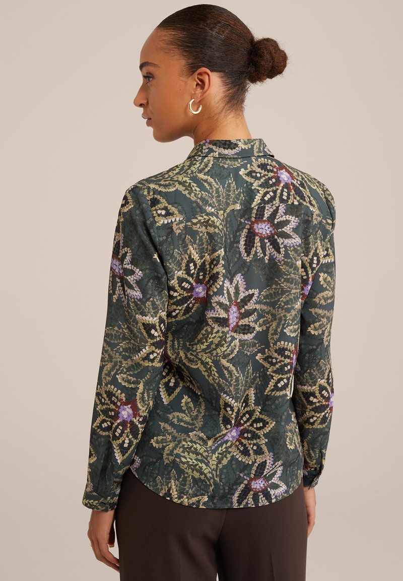 Lange mouwblouse in donkergroene stof met een bloemenpatroon in paarse en crèmekleurige accenten. Ronde zoom en subtiele textuurdetails.