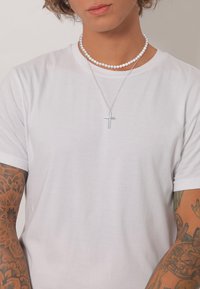 Weißes T-Shirt aus Baumwolle mit kurzen Ärmeln, verziert mit einer silbernen Kreuzkette und einem Perlenchoker, mit detaillierten Tätowierungen am Arm.