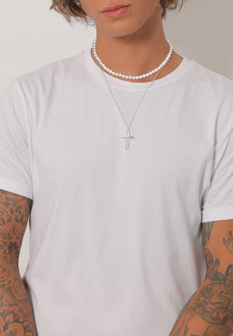 Weißes T-Shirt aus Baumwolle mit kurzen Ärmeln, verziert mit einer silbernen Kreuzkette und einem Perlenchoker, mit detaillierten Tätowierungen am Arm.