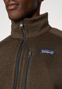 Brown fleece jacket with a high collar, zippered front, and dual zip pockets. Features blue and red Patagonia logo on chest. Textured fabric.