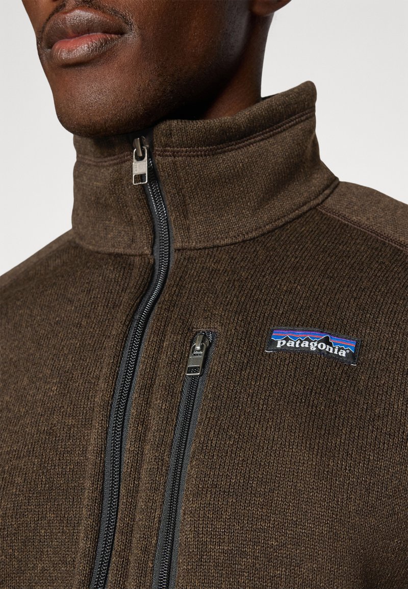 Veste en polaire marron avec un col montant, une fermeture éclair à l'avant et deux poches zippées. Présente un logo Patagonia bleu et rouge sur la poitrine. Tissu texturé.
