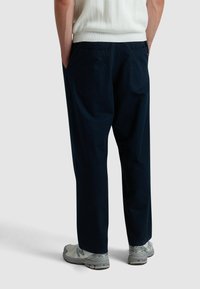 Donkerblauwe katoenen broek met een relaxte pasvorm, voorzien van twee achterzakken en gestyled met grijze sportschoenen.
