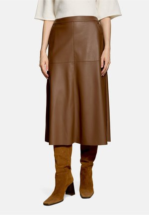 Femme portant une jupe midi en cuir marron associée à des bottes en daim marron montant jusqu’aux genoux et un haut blanc à manches longues, debout, les mains détendues.