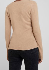 Maglione beige a maglia con un design aderente e maniche lunghe, caratterizzato da un leggero scollo sul retro e un tessuto morbido e texture. Abbinato a jeans scuri.