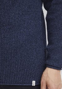 Suéter de punto azul marino con un tejido texturizado, que presenta puños acanalados y una etiqueta en el bajo. Diseño sencillo y casual.