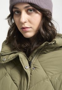 Chaqueta acolchada verde oliva con cuello alto, cierre de cremallera, botones a presión y un sutil detalle de logo. Combinada con un gorro de punto de color púrpura claro.