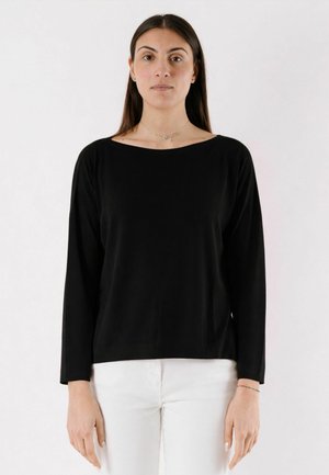 Donna con capelli lunghi e scuri che indossa una maglia nera a maniche lunghe e pantaloni bianchi, in piedi davanti a uno sfondo bianco uniforme.