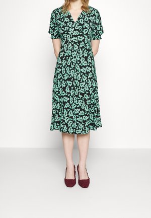 Woman wearing a black midi dress with green abstract pattern, short ruched sleeves, and burgundy woven heels, standing against a white background.