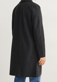 Cappotto in lana carbone con taglio su misura, maniche lunghe e colletto a punta, caratterizzato da una texture liscia e un design pulito e minimalista.
