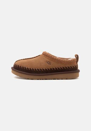 Καφέ σουέντ παντόφλα UGG τύπου slip-on με παχιά σόλα, ορατές ραφές, πλεκτό γιακά και ταινία έλξης στη φτέρνα, εμφανίζεται σε προφίλ σε λευκό φόντο.