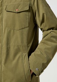 Oliwkowa kurtka o gładkiej fakturze, z kieszenią na guziki na piersi, kieszeniami bocznymi oraz zapięciem na napy na mankietach dla pewnego zamknięcia.