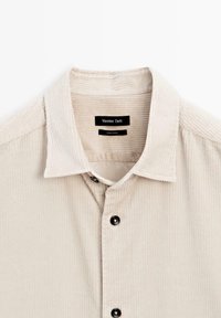 Beige kordflorskjorta med spetsig krage, svarta knappar och diskreta vertikala ränder. Etiketten visar "Massimo Dutti" och skötselinformation.