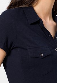 Žena oblečená v tmavě modré krátkosrsté košili s límečkem, zapínáním na knoflíky a náprsní kapsou, zobrazující horní část trupu a krk.