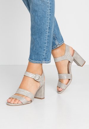 Sandales à talons grises avec une texture reptile, dotées de larges brides, d'une boucle à la cheville et d'un talon bloc. Portées avec un jean bleu retroussé.