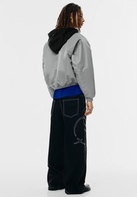 Grijze bomberjack met zwarte capuchon, geribbelde manchetten; gecombineerd met losse zwarte broek met witte stiksels en ingewikkelde grafische ontwerpen.