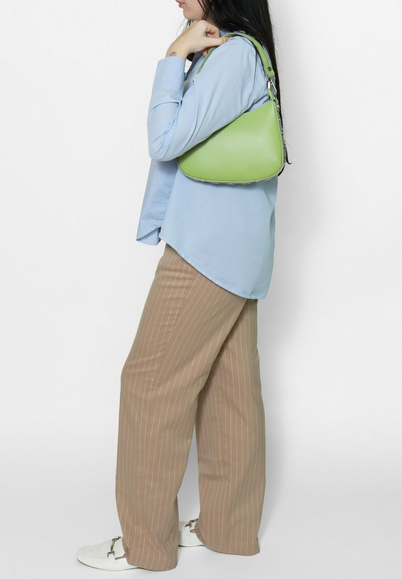 Femme portant une chemise bleu clair et un pantalon beige à fines rayures, avec un petit sac vert porté à l'épaule, sur fond blanc.